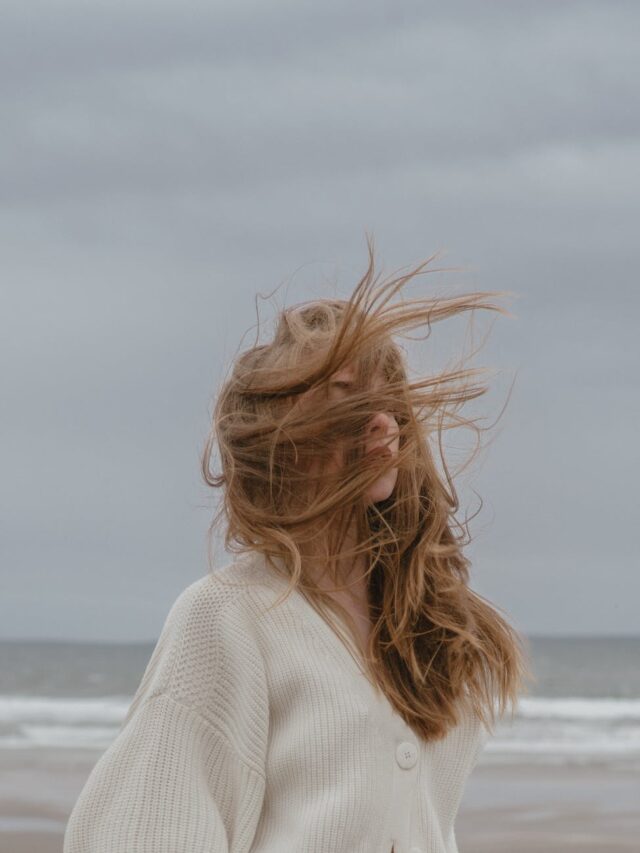 dreamy young woman with windy hair recreating on seashore against cloudy sky