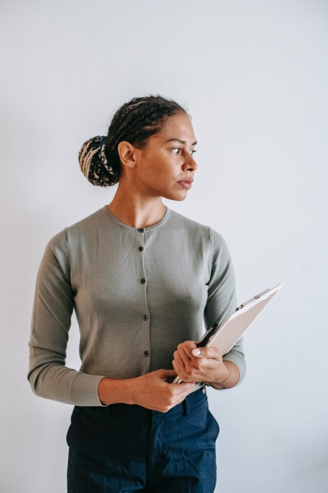 focused ethnic woman with clipboard standing against white wall