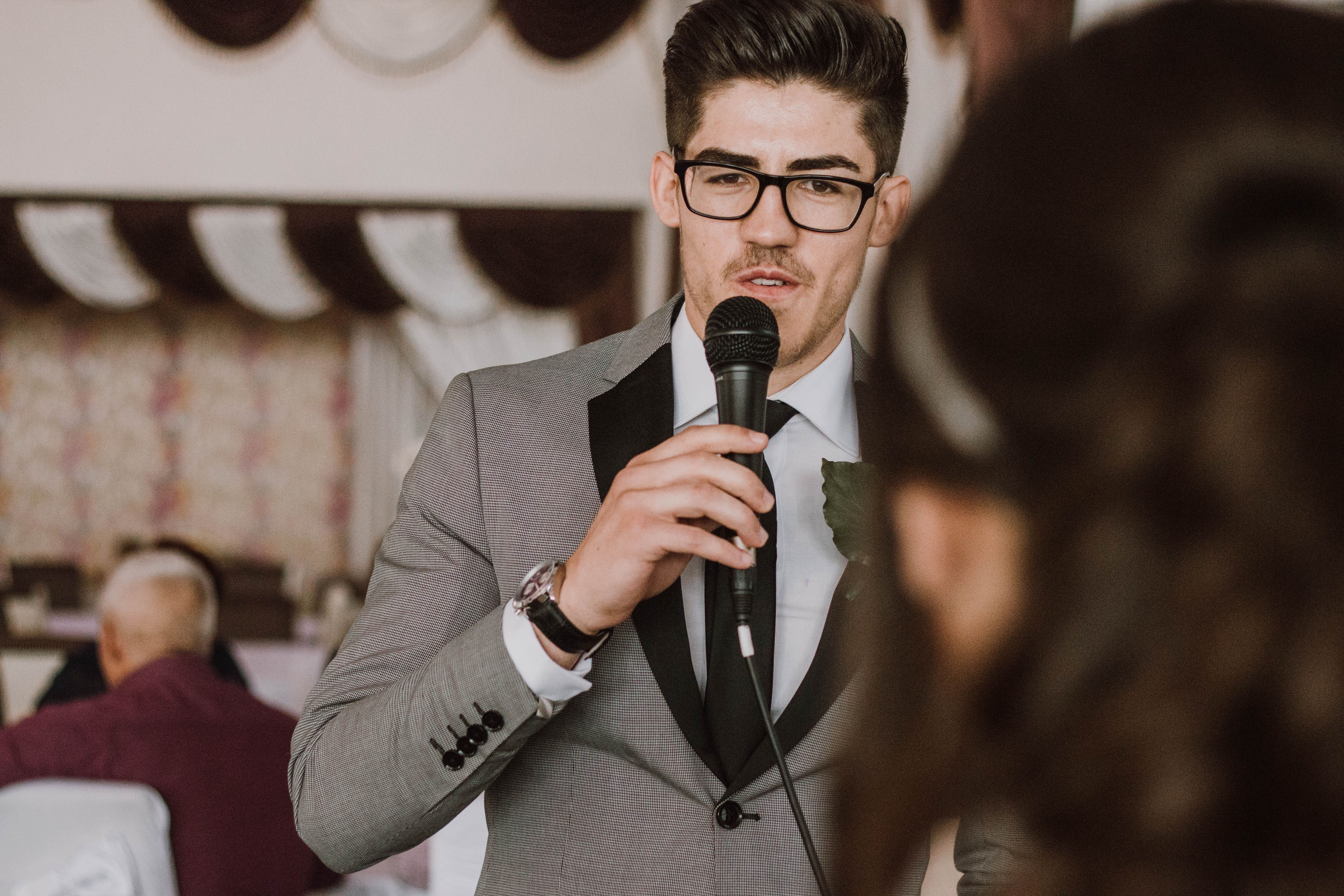 A groomsman in a suit speaking into a microphone to deliver a wedding toast